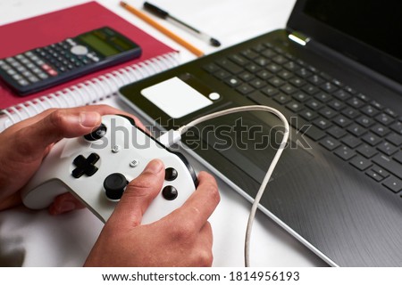 Young man plays a video game with a wired control on his laptop while leaving school aside in times of pandemic, in the background school supplies such as a notebook, calculator, pencil and laptop