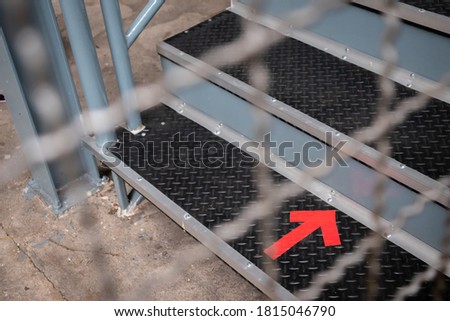 Red forward arrow sign shown on the first step of metal stair in factory with blurred wire mesh fence for restricted area background.