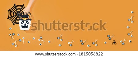Child hand holding Halloween candy bucket face of jack O Lantern with lollypops with spiders and nets on orange background and bloody eyes sweets. Flat lay trick or treat with copy space