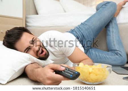 Lazy young man with bowl of chips watching TV while lying on floor at home Royalty-Free Stock Photo #1815140651