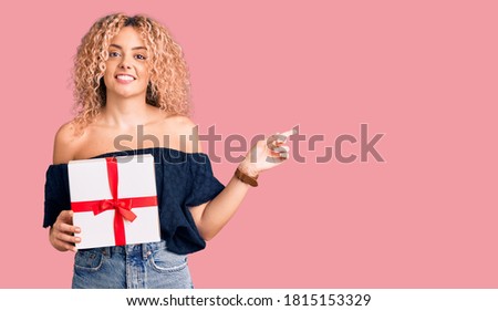Young blonde woman with curly hair holding gift smiling happy pointing with hand and finger to the side 