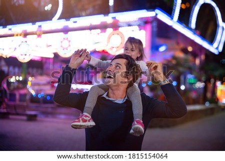 Authentic shot of a happy smiling father carrying his little daughter on a shoulders having fun together in amusement park with luna park lights at night. Royalty-Free Stock Photo #1815154064