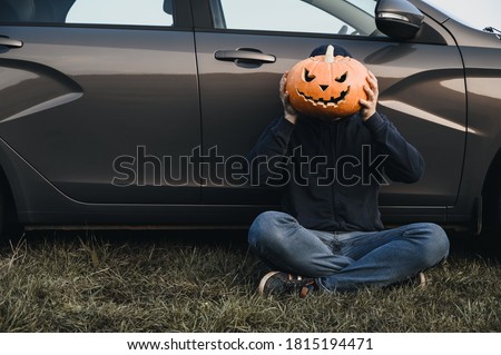 An unrecognizable adult man sits near a car on the grass and holds in front of his face a carved pumpkin for Halloween, outdoors. Copy space. Faceless concept