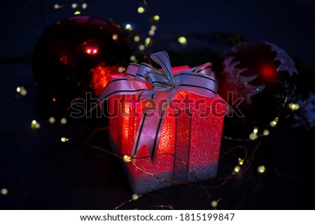 A glowing box with a gift in the dark among the lights of the garland and bright red balls. A picture in a dark key. selective focus.