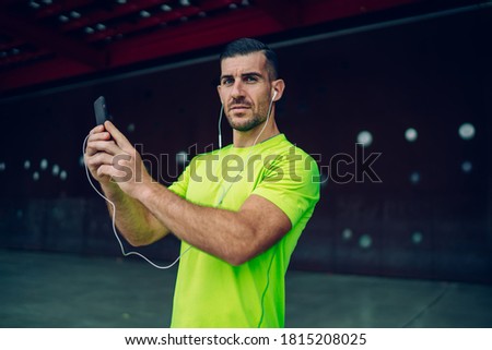 Side view of serious young male athlete in sportswear taking selfie on smartphone while listening to music in earphones during break after training near modern building in city