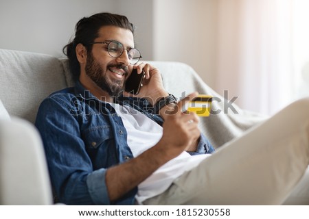 Online Banking. Handsome Indian Man Talking On Cellphone And Using Credit Card And At Home, Paying In Internet, Making Shopping Or Money Transfer, Managing Bills, Sitting On Couch In Living Room Royalty-Free Stock Photo #1815230558