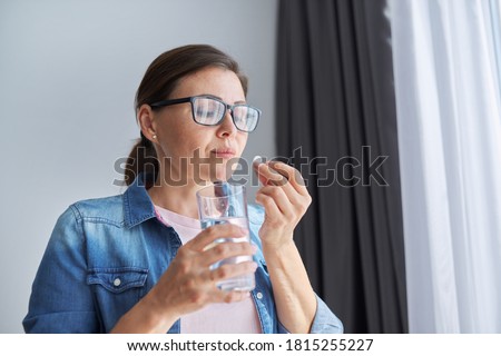 Mature middle-aged woman in casual clothes at home holding pill and glass of fresh water. Headaches, depression, medicine supplements vitamins, sedatives, menopause Royalty-Free Stock Photo #1815255227