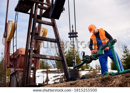 Oil worker in blue overalls, orange vest and helmet using industrial wrench while repairing pipe of oil well pump jack, tightening bolt on pipe. Concept of petroleum industry and repair.
