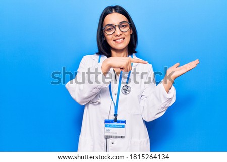 Young beautiful latin woman wearing doctor stethoscope and id card amazed and smiling to the camera while presenting with hand and pointing with finger. 