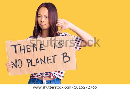 Young beautiful chinese girl holding there is no planet b banner with angry face, negative sign showing dislike with thumbs down, rejection concept 