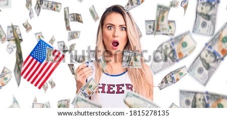 Young beautiful blonde woman wearing cheerleader uniform holding united states flag scared and amazed with open mouth for surprise, disbelief face