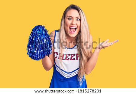 Young beautiful blonde woman wearing cheerleader uniform holding pompom celebrating victory with happy smile and winner expression with raised hands 