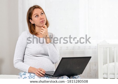 A pregnant woman is sitting on the bed with a thoughtful face, looking up and using a laptop. White room in the background. Concept of modern technologies and freelancing. Copy space