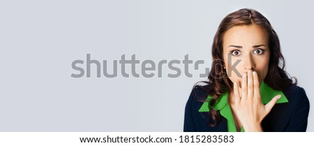 Surprised excited young businesswoman in confident black suit and green blouse, covering with hand her mouth, copy space place for some text, advertising or slogan, over grey background. Studio photo.