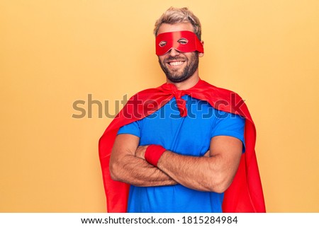 Handsome blond man wearing super hero costume with mask and cape over yellow background happy face smiling with crossed arms looking at the camera. Positive person.