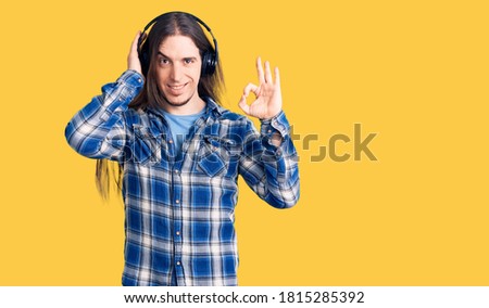 Young adult man with long hair listening to music using headphones doing ok sign with fingers, smiling friendly gesturing excellent symbol 