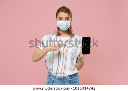 Young blonde woman in casual shirt sterile face mask to safe from coronavirus virus covid-19 during pandemic quarantine hold mobile phone with black blank screen isolated on pastel pink background. 