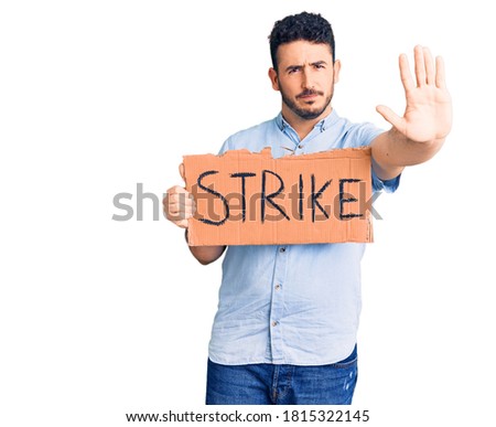 Young hispanic man holding strike banner cardboard with open hand doing stop sign with serious and confident expression, defense gesture 