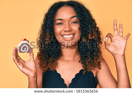 Young african american woman holding cupcake doing ok sign with fingers, smiling friendly gesturing excellent symbol 