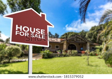 A red house shaped For Sale sign in front of a bungalow with a large lawn. Real estate concept.