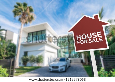 A red house shaped For Sale sign in front of a white modern house. Real estate concept.
