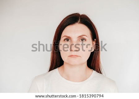Portrait of a serious woman dressed in casual white t shirt, has minimal makeup, poses indoor on a white background. Copy, empty space for text