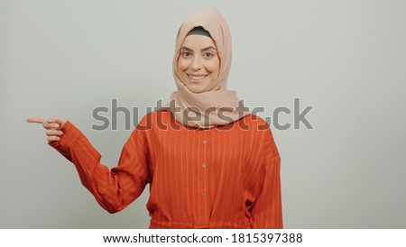 Young Muslim woman wearing a hijab points to the left of the screen with one finger on a white background.White background.