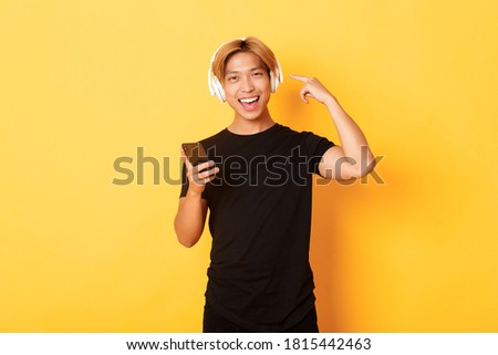 Happy satisfied asian guy likes music or podcast, pointing finger at headphones, holding smartphone, standing yellow background