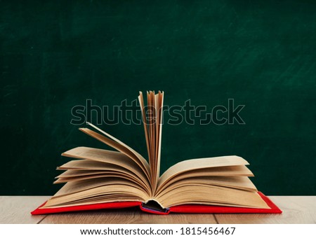 Open book textbook in a red cover on a wooden desk on the background of a green school chalkboard. Back to school distance home education.Quarantine concept of stay home