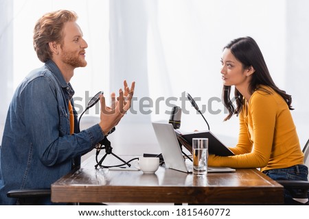 side view of redhead man gesturing while talking to young asian radio host in studio