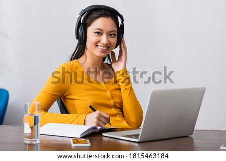 young asian telewoker touching chin and holding pen while sitting at laptop and looking at camera