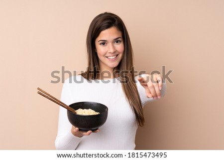 Teenager girl over isolated background points finger at you with a confident expression while holding a bowl of noodles with chopsticks