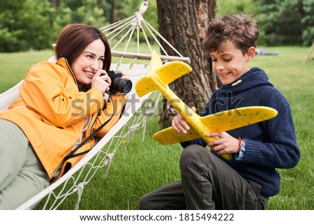 Cheerful young woman using camera and smiling while adorable boy playing with toy airplane
