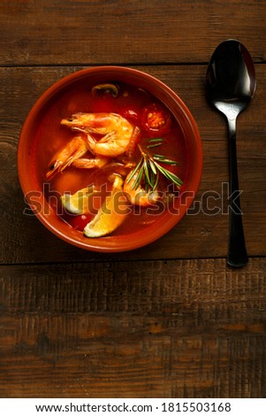 Tom yam soup with shrimp and lime in a brown plate on a wooden table next to a spoon.