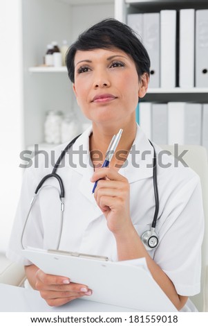 Thoughtful female doctor writing on clipboard at medical office