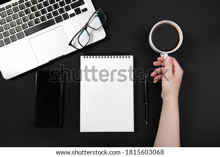  hand holds a cup of coffee on a working black table next to a laptop and accessories. notepad, headphones, pen. Top view with copy space for text input. High quality photo