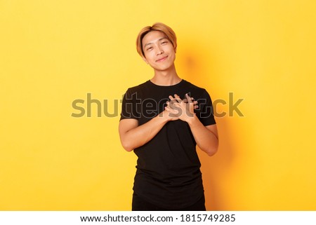 Portrait of touched handsome asian guy holding hands on heart and smiling flattered, yellow background Royalty-Free Stock Photo #1815749285