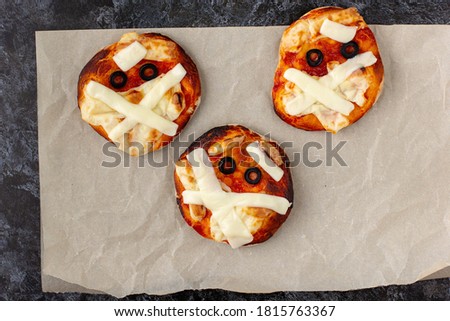 Mini pizza as mummy for kids with cheese, olives and ketchup. Funny crazy Halloween food for children on dark background