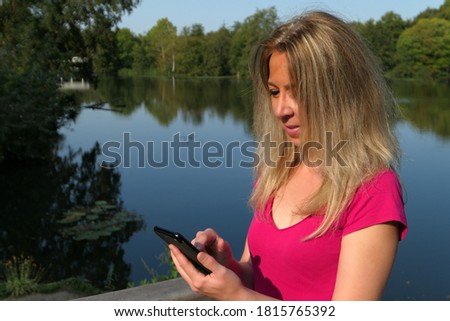 Attractive blonde woman in a park looking at her smartphone. Concept of online shopping on the internet. Use of technology to communicate. Deliberately blurred background.