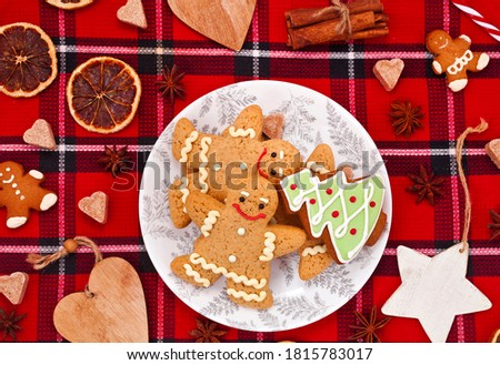 Christmas greeting card. Gingerbread men with fir shaped cookie in a plate, sugar hearts, dried orange slices and spices on a red fabric background.