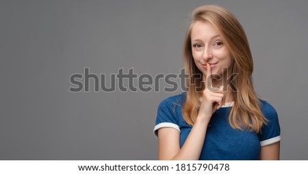 Pretty young blonde woman, dressed in blue t shirt, keeps finger on lips, making hush gesture on gray background. Shh, silence concept. Copy space for your text