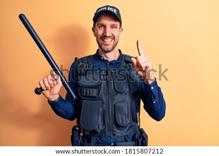 Handsome policeman wearing uniform and bulletproof holding baton over yellow background smiling with an idea or question pointing finger with happy face, number one