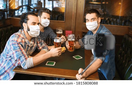 male friendship, leisure and pandemic concept - men or friends in face protective medical masks for protection from virus drinking beer and taking picture with smartphone selfie stick at bar or pub