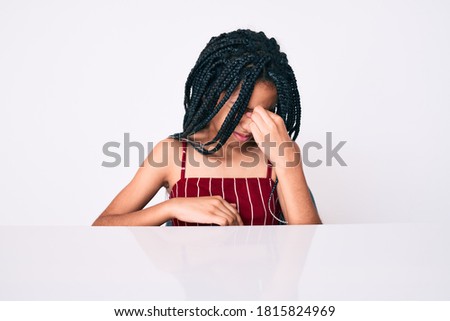 Young african american girl child with braids wearing casual clothes sitting on the table tired rubbing nose and eyes feeling fatigue and headache. stress and frustration concept. 