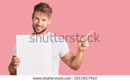 Young caucasian man holding blank empty banner smiling happy pointing with hand and finger to the side 
