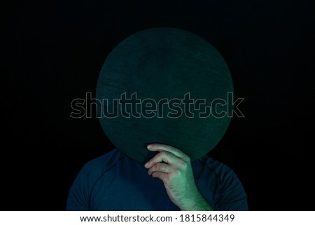 Young guy covered his face with a black round disk in neon light on a black. Minimalism retro style concept.