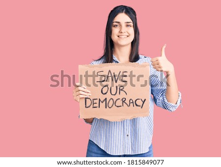 Young beautiful girl holding save our democracy cardboard banner smiling happy and positive, thumb up doing excellent and approval sign 
