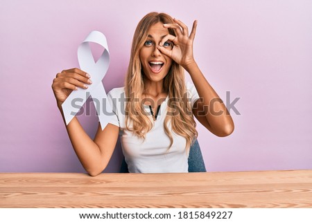 Beautiful blonde young woman holding white ribbon smiling happy doing ok sign with hand on eye looking through fingers 