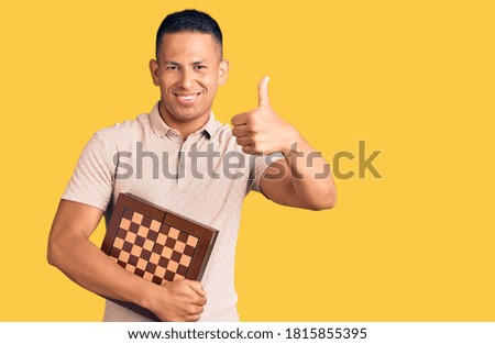 Young handsome latin man holding chess smiling happy and positive, thumb up doing excellent and approval sign 