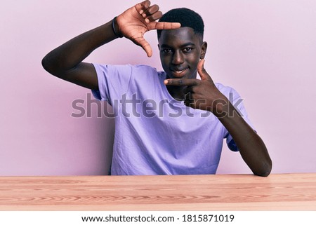 Young african american man wearing casual clothes sitting on the table smiling making frame with hands and fingers with happy face. creativity and photography concept. 